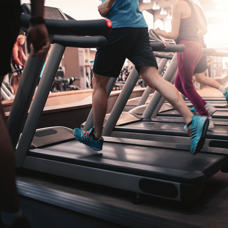 Personnes actives courant sur des tapis de course dans une salle de sport, jambes en mouvement et chaussures de sport bleues.