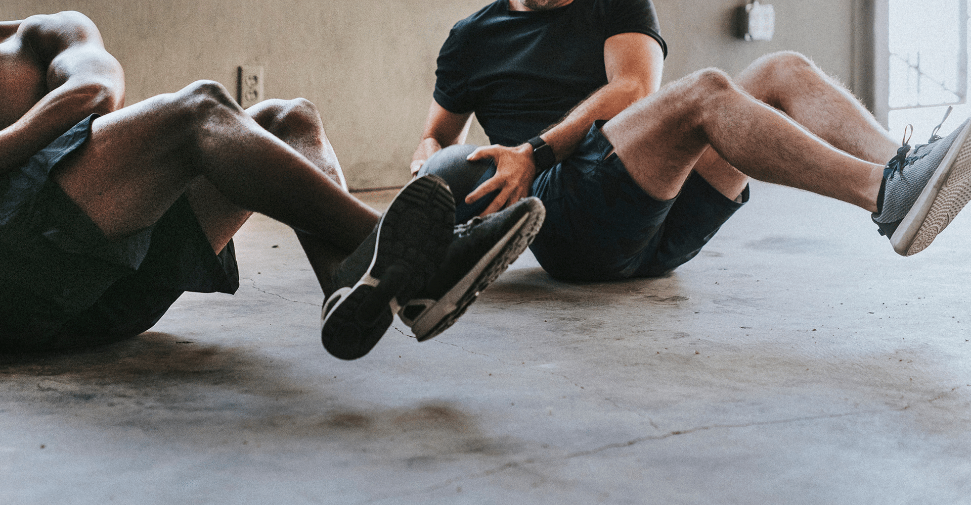 Deux hommes musclés entraînent leurs abdos sur un sol en béton, l'un torse nu, l'autre avec un médecine-ball. Séance intense.