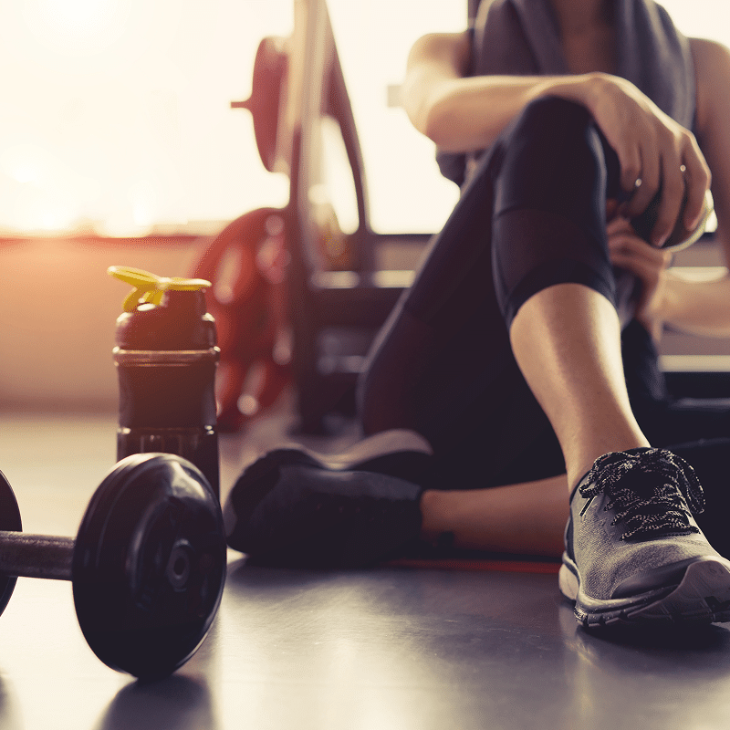 Athlète femme en tenue de sport noire se repose après l'entraînement, avec haltère et gourde au sol dans une salle de sport lumineuse.