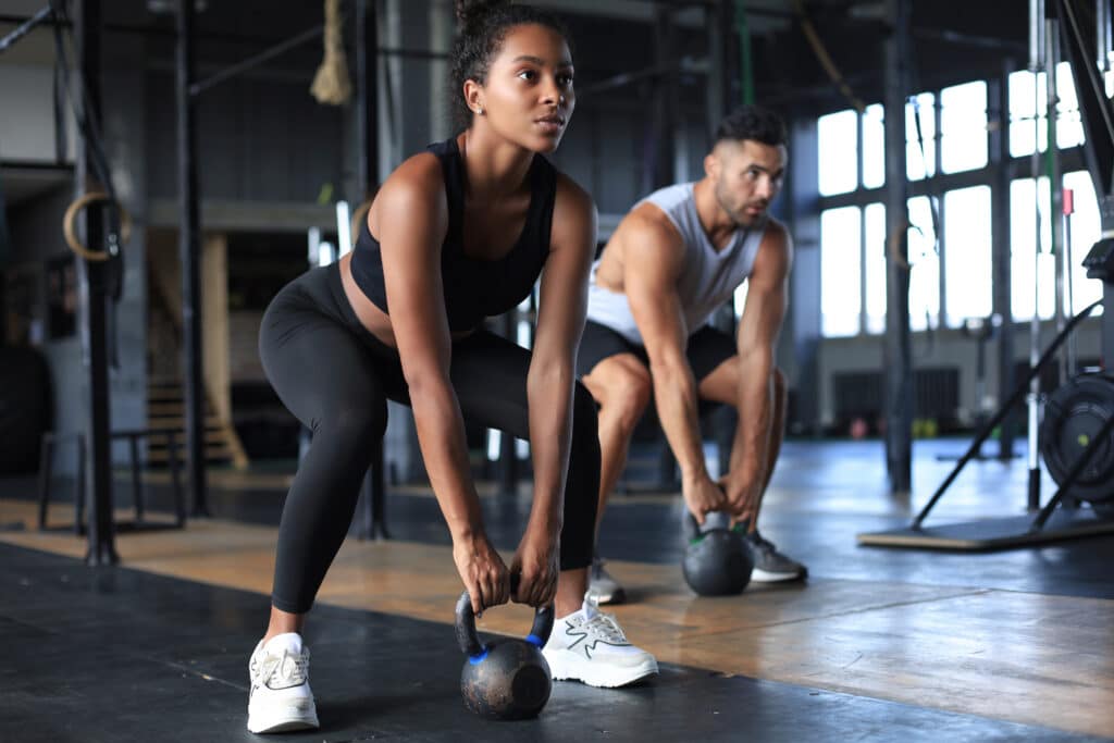 Femme et homme musclés soulevant des kettlebells de 12 kg dans une salle de sport. Ils sont concentrés sur leur entraînement de force.