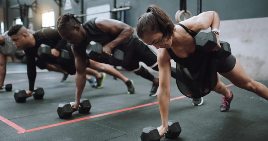 Quatre personnes s'entraînent au renegade row avec haltères 10KG dans une salle de sport. Concentrés sur la musculation.