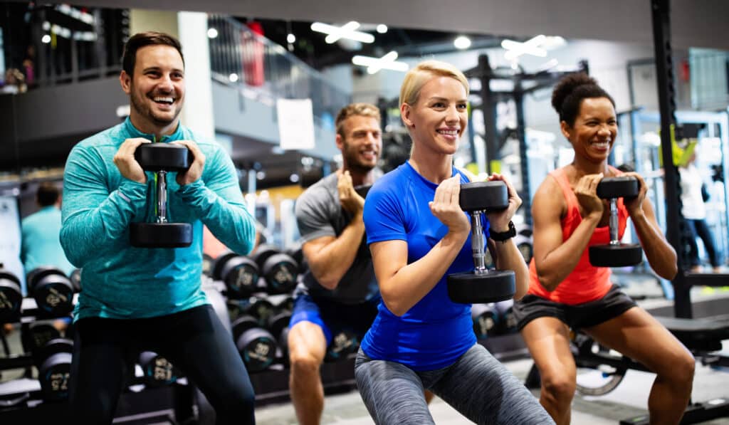 Quatre personnes souriantes s'entraînent avec des haltères dans une salle de sport, effectuant des squats.