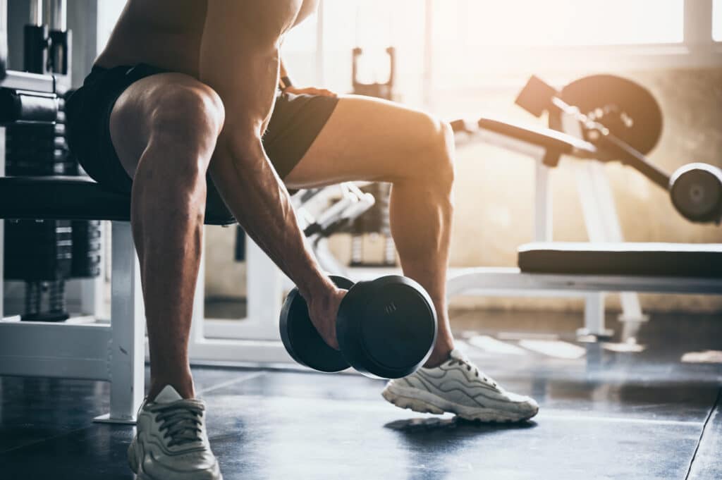 Homme musclé sans chemise assis sur un banc de gym, soulevant un haltère. Lumière chaude et équipement de fitness en arrière-plan.