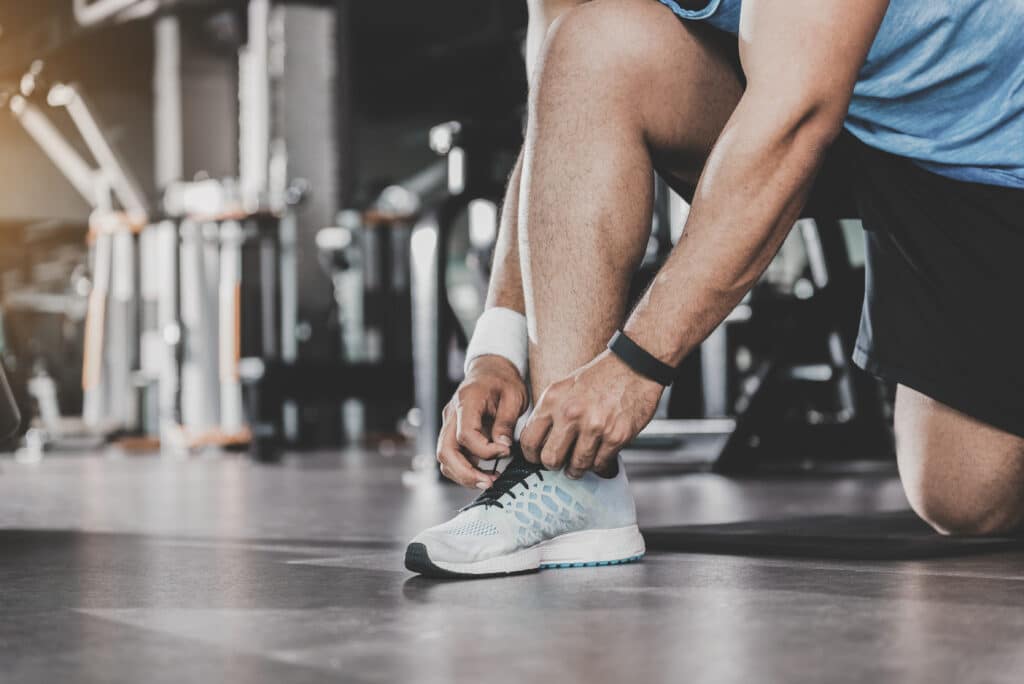 Un homme noue les lacets de ses chaussures de sport bleues et blanches au sol d'une salle de gym, se préparant à l'entraînement.